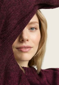 Bordeaux gebreide trui met een textuurpatroon, die gedeeltelijk een gezicht met een lichte huid en blond haar bedekt, tegen een neutrale achtergrond.