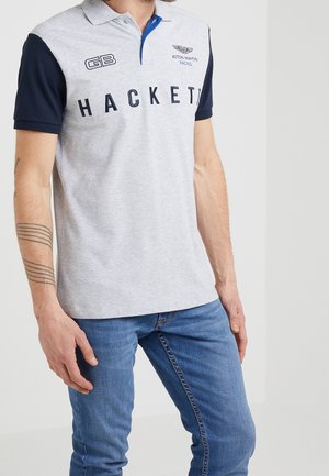 Hombre con camisa polo gris claro con mangas azul marino, logo de Hackett y emblema de Aston Martin Racing, combinado con jeans azules.