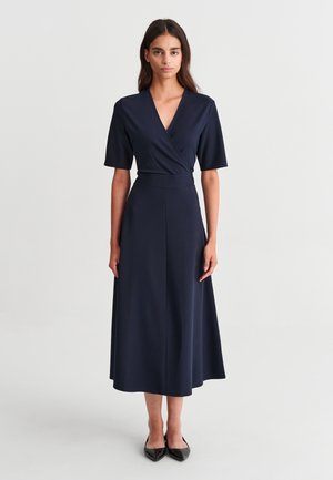 Femme debout portant une robe midi bleu marine à manches courtes, col en V, taille ajustée et chaussures noires à bout pointu sur un fond uni.