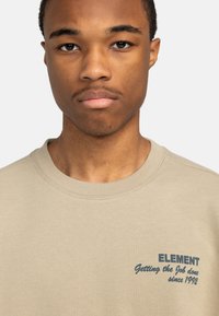 Sudadera beige hecha de un material suave con un cuello redondo. Presenta texto azul oscuro que dice "ELEMENT Haciendo el trabajo desde 1992."