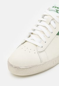 Hvite skinnsneakers med grønne detaljer, flate lisser, perforert tå, teksturert gummisåle, og en lett slitt utseende.