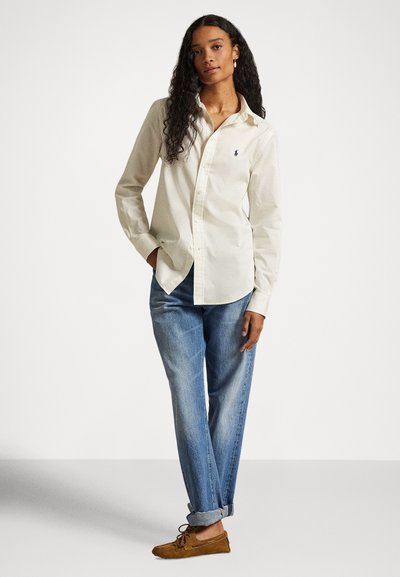 Kvinne med langt krøllete hår iført kremfarget knappeskjorte, oppbrettede blå jeans og brune mokasinsko, stående med den ene hånden i lommen.