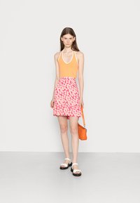 Top halterneck laranja emparelhado com uma saia rufada floral rosa. A modelo usa sandálias brancas e segura uma bolsa laranja. Design simples e contemporâneo.