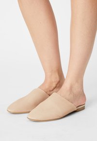 Beige mockasins i mocka med spetsig tå, låg träklack och slät yta, med en öppen häldesign för enkel påtagning.