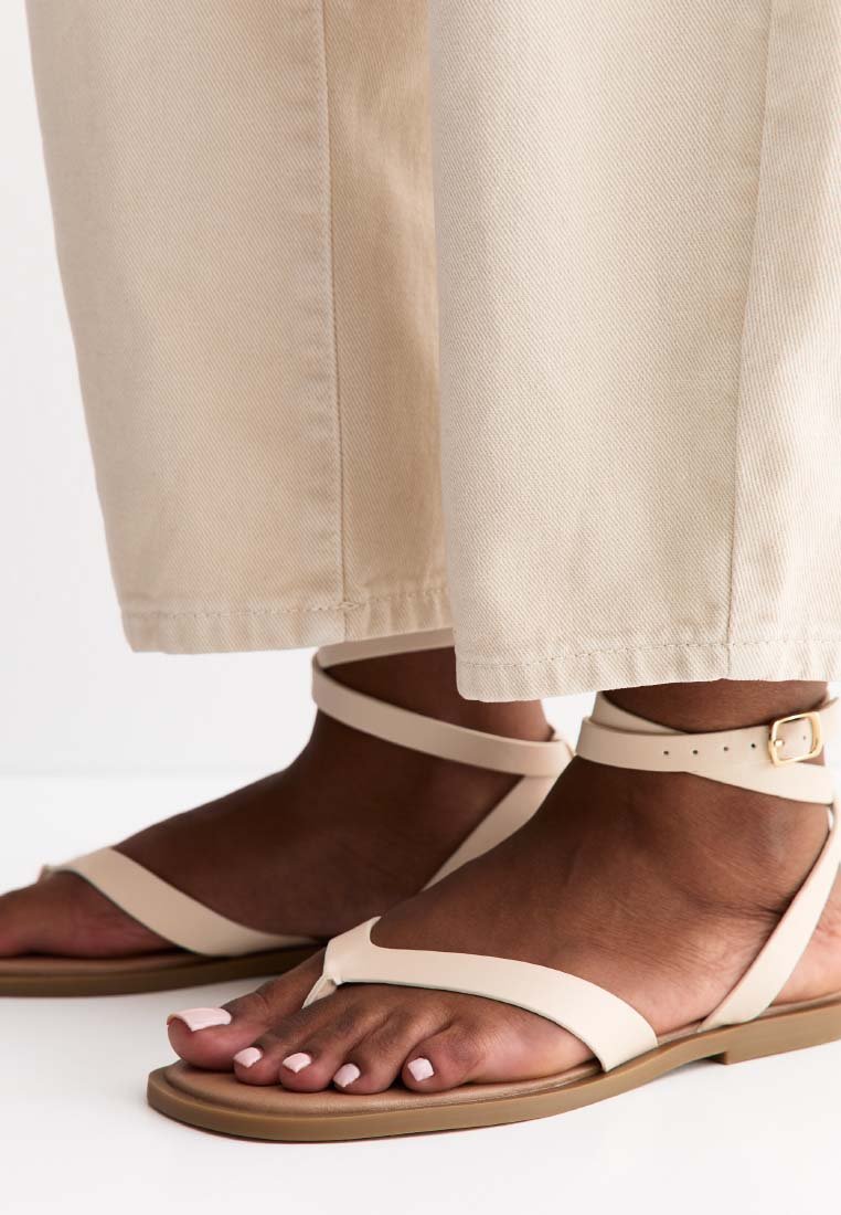 Pieds avec des sandales à bride cheville beige clair et un vernis à ongles rose clair, portant un pantalon beige à jambes larges coupé au-dessus des chevilles.