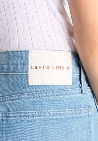 Jeans in denim azzurro chiaro con un'etichetta in pelle rettangolare bianca sulla cintura, impressa con "LEVI'S LINE 8" in lettere color rame.