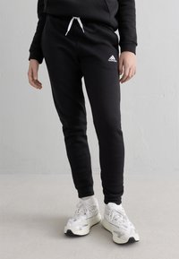 Person som bär svarta Adidas träningsbyxor med vita dragsnören och vita sportskor, stående på en ljusgrå golv mot en vit vägg.