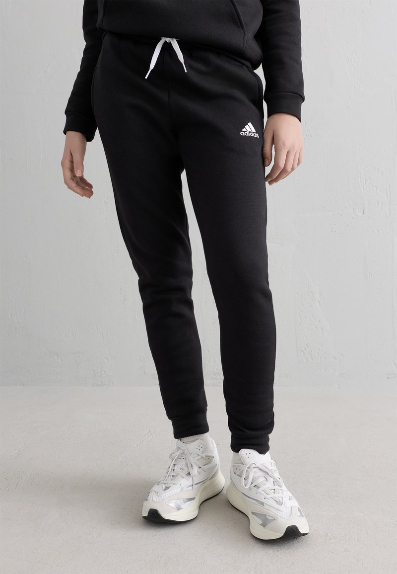 Person som bär svarta Adidas träningsbyxor med vita dragsnören och vita sportskor, stående på en ljusgrå golv mot en vit vägg.