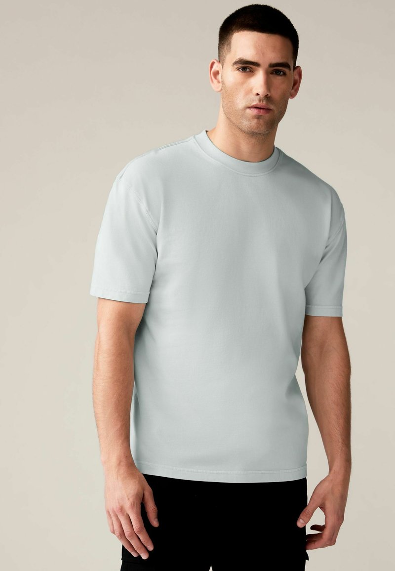 Tricou scurt din bumbac de culoare albastru deschis, cu mânecă scurtă, având un guler rotund și o croială lejeră. Design simplu, fără modele sau accente.