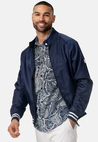 Giacca bomber navy dal texture liscio e lucido, con colletto nero e polsini a strisce bianche, indossata sopra una camicia blu con motivo floreale.