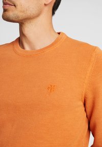 Pull en tricot texturé orange avec un col rond et un logo brodé discret sur la poitrine. Doté d'un col et de bords-côtes.