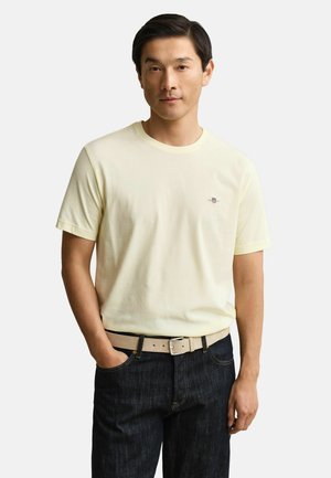 Homme aux cheveux noirs courts portant un t-shirt à manches courtes jaune pâle, ceinture beige et jean bleu foncé, debout avec une main dans la poche.