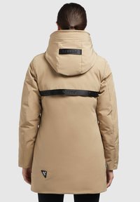 Bēša parka jaka ar augstu apkakli, melnām logo akcentiem un trapezoīda formas zīmola etiķeti kreisajā apakšējā stūrī. Gluda tekstūra, garas piedurknes.