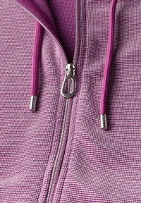 Veste à zip violet, en tissu texturé avec un motif à carreaux, fermeture éclair argentée et cordons roses avec embouts métalliques.
