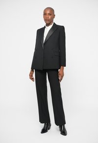 Blazer noir sur mesure avec revers en satin, design croisé, associé à un pantalon noir à jambes droites et des bottines noires élégantes.