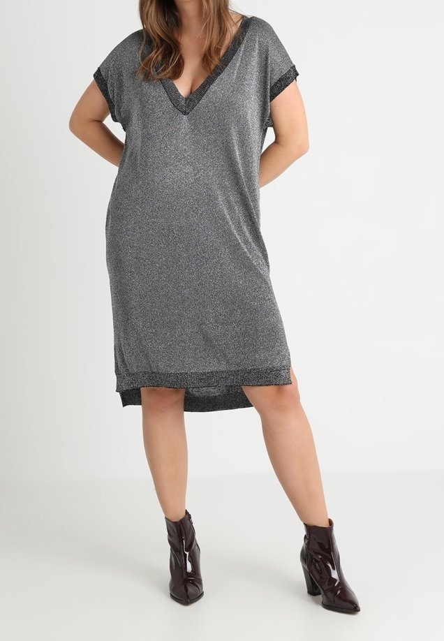 Femme portant une robe grise ample, longueur genoux, avec manches courtes et col en V, associée à des bottines marron foncé brillantes.