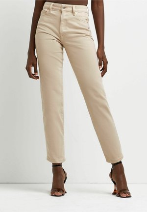 Beige højhvide jeans med slank pasform, båret af en stående person, kombineret med sorte åbne tå ankelrem-sandaler mod en ensfarvet hvid baggrund.