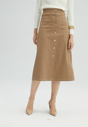 Femme portant une jupe midi taille haute couleur beige avec boutons sur le devant, associée à des talons beiges et un haut côtelé à manches longues crème.