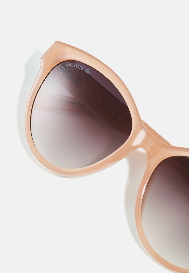 Occhiali da sole cat-eye rosa con lenti scure a gradiente e piccolo logo Lacoste nell'angolo superiore della lente sinistra.