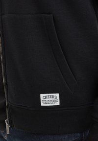 Sudadera con cremallera negra que cuenta con un bolsillo frontal, dobladillo acanalado y una etiqueta blanca que dice "CREEKS" con detalles en texto. Material suave de sudadera.