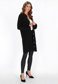 Cardigan nero a maglia con un motivo testurizzato e bottoni, indossato sopra una blusa chiara e abbinato a leggings neri e tacchi.