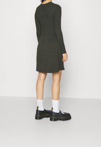 Mörkgrön ribbad stickad klänning med långa ärmar och åtsittande överdel, med en utsvängd kjol och sidoflikar, matchad med svarta skor och vita strumpor.