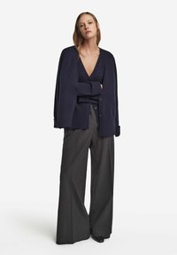 Kvinna står med armarna i kors, iförd marinblå cardigan över en matchande topp och vida mörkgrå byxor mot en vit bakgrund.