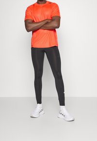 Camiseta deportiva de color naranja brillante con mangas cortas, combinada con leggings ajustados negros y zapatillas blancas. Tejido suave, diseño minimalista.
