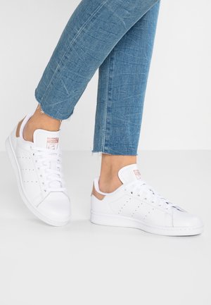 Person iført hvide Adidas Stan Smith sneakers og blå afkortede jeans, der står på en hvid gulv mod en ensfarvet baggrund.