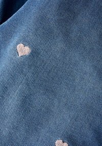 Denim tkanina v umirjenih modrih tonih, ki vsebuje majhne, vezenine roza srčke raztresene po površini. Tekstura se zdi mehka in gladka.