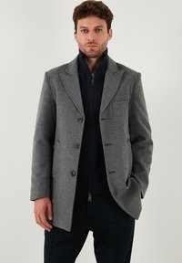 Cappotto grigio in lana con rever a punta, tre bottoni e una tasca sul petto, indossato sopra un maglione blu navy con zip.