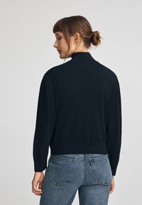 Svart stickad tröja med hög krage och ribbat avslut. Modellen bär den med blå denimjeans, vilket visar en avslappnad, figurnära look.