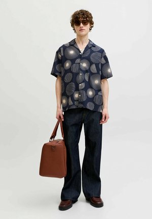Young man wearing sunglasses, navy patterned short-sleeve shirt, dark wide-leg jeans, brown shoes, holding a brown leather bag.