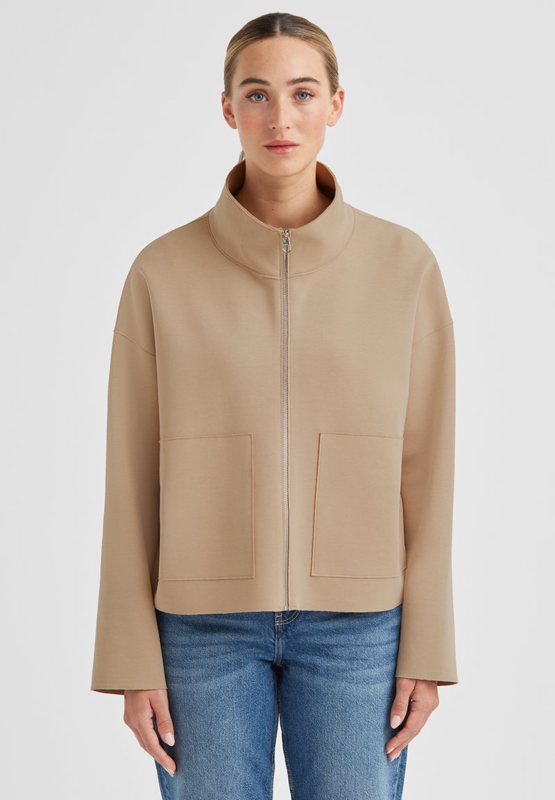 Beige Reißverschlussjacke mit hohem Kragen, zwei Vorder Taschen und weiter Passform. Glatter Stoff mit matter Oberfläche und weiten Ärmeln.