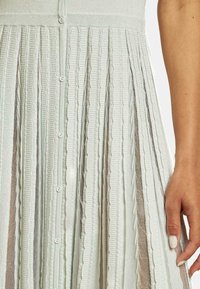Abito lavorato a maglia di colore verde chiaro, con texture a coste, chiusura con bottoni e gonna svasata con pannelli trasparenti alternati.