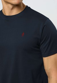 Čovjek u jednobojnoj tamnoplavoj kratkoročavnoj majici s malim crvenim izvezenim logom na lijevoj strani prsa.