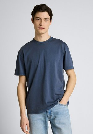 Jonge man in een effen donkerblauw T-shirt en lichtblauwe spijkerbroek, staand met een hand in zijn zak tegen een neutrale achtergrond.