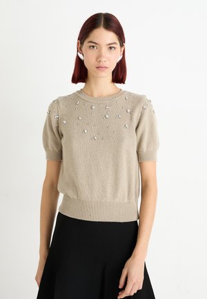 Beige knitted short-sleeve sweater with round neck, featuring scattered clear embellishments and a ribbed hem. Worn with a black skirt.
