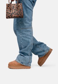 Botas de plataforma de ante marrón, jeans de pierna ancha de color azul claro y un pequeño bolso de mano con estampado de leopardo. Las botas tienen una suela gruesa y una textura suave.