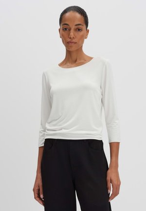 Donna con capelli corti che indossa un top bianco a maniche lunghe e pantaloni neri, in piedi davanti a uno sfondo chiaro uniforme.