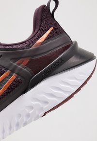 Nike React -juoksukengässä on tummanvioletti verkkopäällinen, metallinkiiltoiset kupariväriset yksityiskohdat ja teksturoitu valkoinen pohja, jossa on viininpunainen pohjalevy.