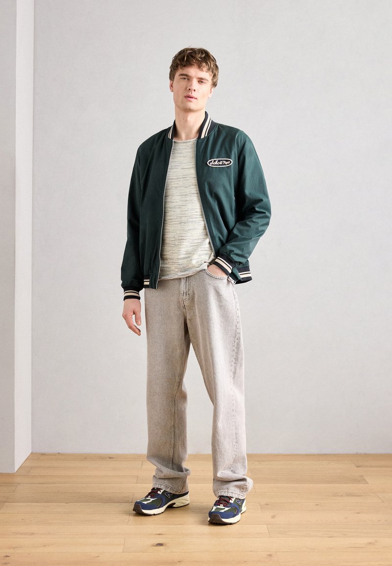 Un jeune homme portant une veste bomber vert foncé, un pull gris clair, un jean beige ample et des baskets multicolores, se tenant sur un sol en bois.