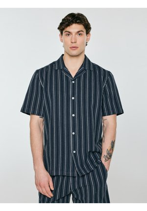 Młody mężczyzna w granatowej koszuli na krótki rękaw z guzikami i dopasowanych spodniach z pionowymi białymi paskami, stoi z jedną ręką w kieszeni.