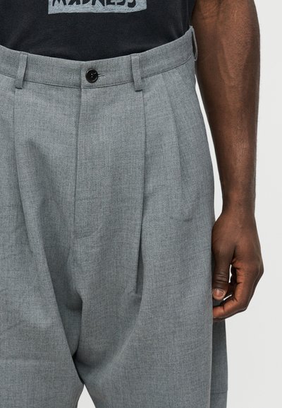 Άνδρας που φοράει γκρι παντελόνι με πιέτες και ψηλή μέση, με θηλιές για ζώνη και μαύρο κουμπί, συνδυασμένο με μαύρο t-shirt με γραφικό σχέδιο.