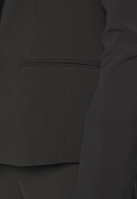 Blazer nero con un tessuto dalla texture liscia. I dettagli degni di nota includono una tasca laterale con un bordo sottile e rever affilati.