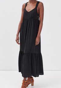 Femme portant une robe maxi noire sans manches avec une jupe à volants et des sandales à talons marron à lanières, debout devant un fond uni.