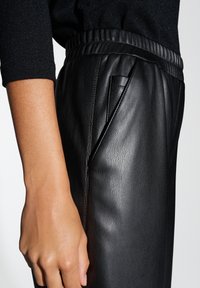 Schwarze Kunstlederhosen mit glatter Textur, elastischem Taillenbund und seitlichen Taschen. Das Design umfasst subtile Nähdetails.