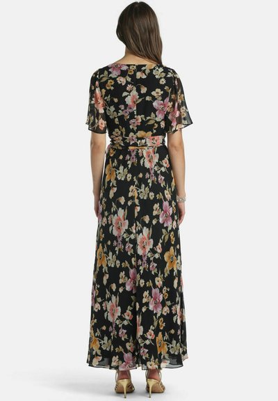 Woman wearing a black floral maxi dress with short sleeves and a belt, standing facing away, wearing tan high-heeled sandals.