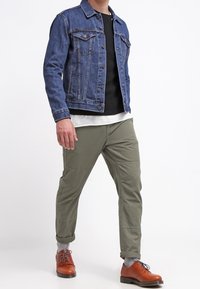 Man som bär en blå denimjacka, svart och vit lagertröja, olivgröna byxor, grå strumpor och bruna läderskor medan han går.