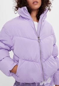 Fioletowa kurtka puchowa z wysokim kołnierzem i zapięciem na zamek. Posiada pikowany wzór i boczne kieszenie. Gładki, lekki materiał.